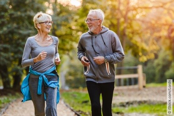 Zwei ältere Personen, eine Frau und ein Mann, joggen lächelnd auf einem Waldweg im Sonnenlicht. Die Frau trägt eine graue Sportjacke und eine blaue Jacke um ihre Hüften, der Mann ist in einen grauen Hoodie gekleidet. Beide tragen Brillen und sehen gesund und aktiv aus, was einen lebensbejahenden Lebensstil und das Engagement für körperliche Fitness und Blasengesundheit im Alter darstellt.