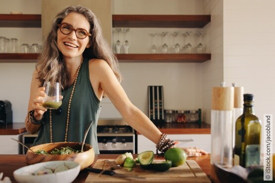 Eine heitere Frau mit grauem Haar und Brille, gekleidet in ein ärmelloses grünes Top, steht in einer hellen Küche und hält ein Glas mit grünem Smoothie. Auf dem Küchentisch sind frische Zutaten wie Avocados, Limetten und eine Schale mit Salat zu sehen, was auf eine gesunde Ernährung hinweist. Die Frau lächelt und scheint eine ausgewogene Lebensweise zu genießen, die auch zur Unterstützung der Blasengesundheit beitragen kann.