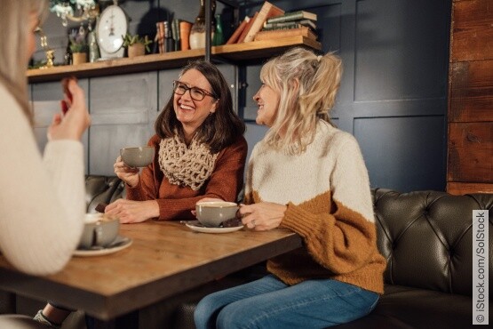 Zwei Frauen mittleren Alters genießen lachend eine Kaffeepause in einem gemütlichen Café mit rustikalem Dekor. Die Frau in der Nahaufnahme hält eine große graue Tasse und trägt eine orange Strickjacke sowie einen braunen Schal, die andere Frau einen cremefarbenen Pullover mit braunen Akzenten. Sie sitzen an einem hölzernen Tisch, und das Ambiente ist warm und einladend, was auf entspannte Gespräche und eine angenehme Auszeit hindeutet.