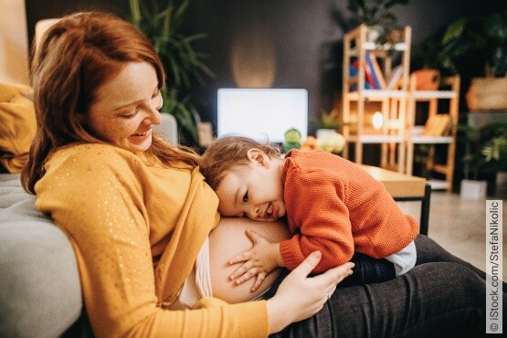 Ein kleines Kind in einem orangefarbenen Pullover schmiegt sich liebevoll an ihre lächelnde schwangere Mama in einem gelben Oberteil. Die Mama umarmt das Kind sanft, während sie auf einem grauen Sofa sitzen, mit einer wohnlichen und pflanzenreichen Einrichtung im Hintergrund.