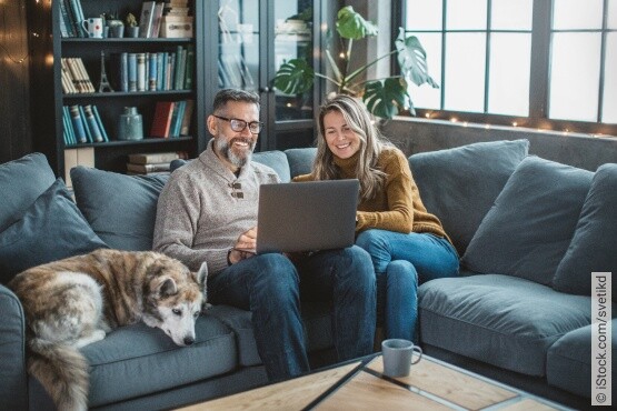 Ein lächelndes Paar mittleren Alters sitzt entspannt auf einem dunkelblauen Sofa in einem gemütlich eingerichteten Wohnzimmer mit Bücherregalen und Pflanzen. Der Mann trägt eine graue Strickjacke und Brille, während die Frau in einem senfgelben Pullover und Jeans lächelt und ein Laptop auf ihren Knien hat. Ein großer Husky liegt neben ihnen auf dem Sofa. Die familiäre Szene drückt Geborgenheit und Wohlbefinden aus, was zur allgemeinen Gesundheit und speziell zur Blasengesundheit beitragen kann.