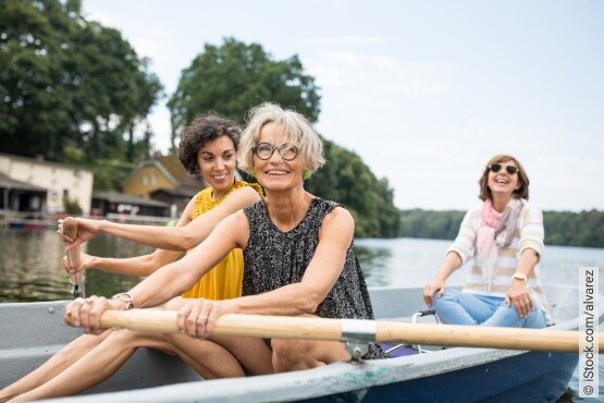 Drei lächelnde Frauen unterschiedlichen Alters genießen eine gemeinsame Ruderbootfahrt auf einem ruhigen See. Die ältere Dame mit grauem Haar und einer schwarzen Tunika sitzt vorne, während die mittlere Frau in einem gelben Top unterstützend hinter ihr steht. Eine weitere Frau mit Sonnenbrille und einem hellen Schal lacht im Hintergrund. Die Szene symbolisiert aktive Freizeitgestaltung und das Teilen von freudvollen Momenten, was zur allgemeinen Gesundheit und zur Unterstützung der Blasengesundheit beiträgt